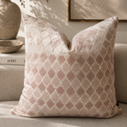 A square throw pillow with a dusty pink and cream geometric trellis pattern, styled on a beige sofa in a warm, sunlit room with a vase of flowers and framed wall art in the background.