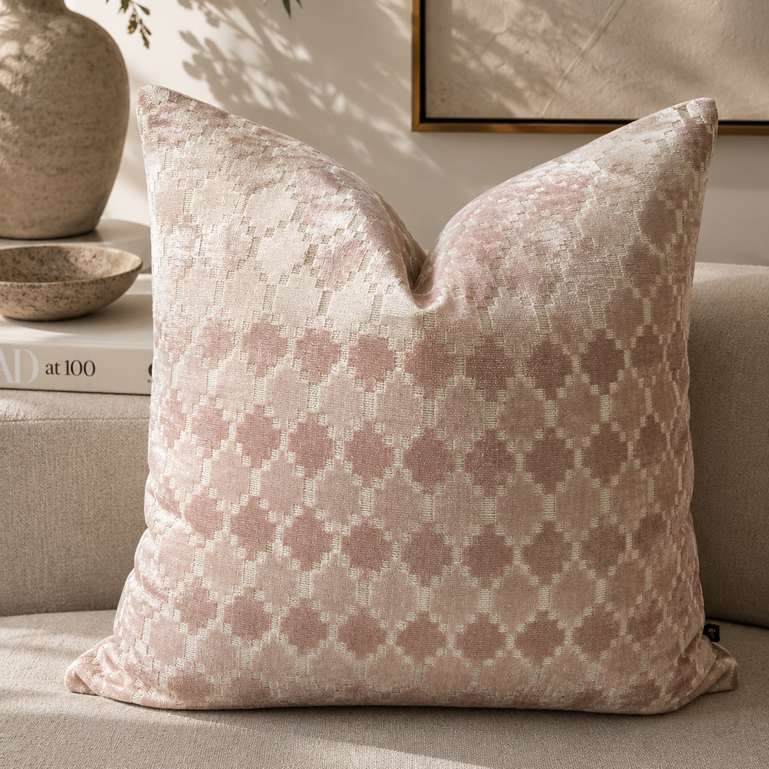 A square throw pillow with a dusty pink and cream geometric trellis pattern, styled on a beige sofa in a warm, sunlit room with a vase of flowers and framed wall art in the background.